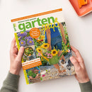 Gartenmagazin „garten spaß“ in den Händen einer Person, schräg auf weißer Fläche, mit grün-gelbem Logo und orangefarbener Gestaltung. Sichtbare Texte: „Das Garten-Magazin mit vielen Schritt-für-Schritt-Anleitungen“, „Gärtnern leicht gemacht“, „Trends für Topf & Terrasse“ und „Volle Kanne Spätsommer“; Titelbild mit einer weiblichen Person im Garten, Sonnenblumen, Gießkanne, Katze und weiteren Pflanzen.