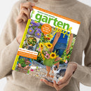 Eine Person im beigefarbenen Strickpullover hält ein schräg ausgerichtetes Gartenmagazin mit grünem Titel „garten“ und gelber Aufschrift „pass“. Das Cover zeigt eine gärtnernde Frau mit grüner Gießkanne und Blumen; lesbare Texte: „September 2026“, „Gärtnern leicht gemacht“, „Blühende Astern in allen Farben“, „Mal anders kochen mit Asia-Blattgemüse“, „Schick & Schnell Pflanzen-Deko im Korb“ und „Volle Kanne Spätsommer“.