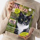Weibliche Person im beigen Strickpullover hält schräg eine Zeitschrift mit grünem Hintergrund und großem Titel „Geliebte KATZE“. Das Cover zeigt eine schwarz-weiße Katze, ein Katzenjunges und eine getigerte Katze sowie Texte wie „Aktuelle Forschung: Wie Katzen uns gesünder machen“, „Experten-Tipps“, „Charakterkopf“ und „Zum Weltkatzentag“.