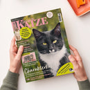 Aufgeschlagenes Titelbild der Zeitschrift „Geliebte KATZE“ mit einer schwarz-weißen Katze im Großformat und zwei kleineren Katzenabbildungen. Sichtbare Texte: „Europas beliebtestes Katzenmagazin“, „Aktuelle Forschung: Wie Katzen uns gesünder machen“, „Charakterkopf“ und „Zum Weltkatzentag“; zwei Hände mit grauen Ärmeln halten das Magazin vor weißem Hintergrund.