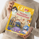 Eine Person im beigefarbenen Strickpullover hält ein schräg fotografiertes GEOlino-mini-Magazin mit gelbem Cover in beiden Händen. Sichtbar sind die Texte „Die Welt für junge Entdeckerinnen und Entdecker“, „ab 5 Jahren“, „Tierische Freundschaft“, „Kinder: Manuela lebt im Regenwald“, „Basteln: Lass die bunten Puppen tanzen“ und „Wissen: Hoch hinaus: So entstehen Berge“ sowie zwei Ferkel, ein Bergfoto und eine gezeichnete Raubkatze.