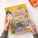 Gelbes GEOlino-mini-Magazin mit dem Titel „GEO lino mini“ und den Aufschriften „Die Welt für junge Entdeckerinnen und Entdecker“, „ab 5 Jahren“ und „Tierische Freundschaft“. Das Cover zeigt zwei Ferkel, eine Kinder-Abbildung, einen gezeichneten Leoparden sowie Textfelder „Kinder: Manuela lebt im Regenwald“, „Wissen: Hoch hinaus – so entstehen Berge“ und „Basteln: Lass die bunten Puppen tanzen“; zwei Hände einer nicht näher bestimmbaren Person halten das Heft.