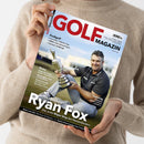 Hände einer Person halten ein schräg fotografiertes Golfmagazin mit beigem Strickpullover im Hintergrund. Das Cover trägt den Titel „GOLF MAGAZIN“, zeigt einen männlichen Golfer mit silbernem Pokal und enthält die Texte „Profigolf“, „The Open 2026“, „Ryan Fox – Der erste Major-Sieg des Neuseeländers“, „Reise“ und „Training“.