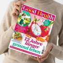 Eine Person in einem beigefarbenen Strickpullover hält ein schräg fotografiertes, glänzendes Magazin aus Papier mit beiden Händen. Das pink-weiße Cover von „Good Health“ zeigt Drachenfrucht, Sanddornbeeren, ein Gemüsegericht und eine springende Frau; sichtbar sind unter anderem „12 Jahre länger gesund leben“, „Endlich schmerzfrei!“ und „Japan-Workout“.
