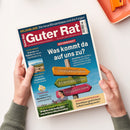 Schräg fotografierte Zeitschrift „Guter Rat“ mit blau-rotem Cover, weißem Rand und zwei Händen am unteren Rand. Sichtbare Texte: „GELDANLAGE – Die neue Börsenblase und die Folgen“, „Reformpaket“, „Was kommt da auf uns zu?“, „Steuerreform“, „Rentenreform“, „Pflegereform“ sowie Beiträge zu Digitalwährung, Urlaubsielen, Eiscreme, Heckenscheren und Wärmepumpenförderung; oben rechts liegen ein orangefarbenes Tablett und ein orangefarbener Gegenstand.