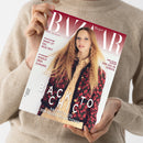 Person im hellbeigen Strickpullover hält ein schräg ausgerichtetes „Harper’s BAZAAR“-Magazin mit rotem und weißem Cover. Darauf eine Frau mit langem welligem Haar und floral gemusterter Jacke sowie die Texte „08/26“, „harpersbazaar.de“, „Smart ist das neue sexy“, „Rosalia über Euphorie“, „Eine Ode an Venedig“, „BACK TO CHIC“ und „Das Beste aus den neuen Kollektionen“.