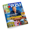 Schräg fotografiertes HÖRZU-Titelbild mit blau-grünem Hintergrund und der Aufschrift „TV-Programm vom 22. bis 28. August 2026“. In der Mitte ein grauhaariger Mann im dunkelblauen Hemd, darunter Texte wie „Deutschland sucht das SUPERHIRN“ und „Neue Quizshow mit Jörg Pilawa“; außerdem eine Frau, ein Puma, eine Weinlandschaft mit Kirche sowie Hinweise auf „Marianne Koch wird 95“, „Zauberhaftes Elsass“ und „Im Reich der Berglöwen“.