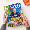 Auf blau-grünem HÖRZU-Magazincover hält eine Person mit beiden Händen eine Ausgabe vor weißem Hintergrund; darauf ein lächelnder Mann im blauen Hemd. Sichtbare Texte: „TV-Programm vom 22. bis 28. August 2026“, „Der große Bundesliga-Spielplan“, „Deutschland sucht das SUPERHIRN“, „Neue Quizshow mit Jörg Pilawa“ und „Im Reich der Berglöwen“; oben rechts liegt ein orangefarbenes Tablett.
