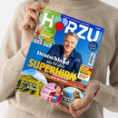 Person in beigem Strickpullover hält ein schräg ins Bild gedrehtes, blau-grünes TV-Magazin mit dem Titel „HÖRZU“. Auf dem Cover stehen „Ihr Premium TV-Programm vom 22. bis 28. August 2026“, „Deutschland sucht das SUPERHIRN“, „Neue Quizshow mit Jörg Pilawa“, „Zauberhaftes Elsass“, „Marianne Koch wird 95“ und „Im Reich der Berglöwen“; abgebildet sind ein Mann, eine Frau, eine Landschaft und ein Puma.
