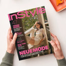 Hochformatiges, leicht gedrehtes Modemagazin mit schwarzem Cover und pinkem „InStyle“-Schriftzug, von zwei Händen gehalten. Abgebildet ist eine weibliche Person in hellem Outfit am Fenster; sichtbare Texte: „47 TREND-SHOES“, „Micro-Shorts – Herbst?!“, „Leslie Mann“, „SKIN-TONE HAIR“ und „NEUE MODE“.