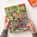 Aufgeschlagene Zeitschrift „LandIDEE“ in zwei Händen mit grauen Strickärmeln auf weißem Untergrund; Titelbild mit blühendem Garten, Obst und herbstlicher Dekoration. Sichtbare Texte: „LandIDEE“, „September & Oktober 2026“, „137 Ideen zum Selbermachen“, „Land erleben und genießen“, „Leckere Rezepte mit Äpfeln und Quitten“, „Herbstliche Deko für den Balkon“ und „Leuchtfeuer für die Sinne“; oben rechts liegt ein orangefarbenes Tablett.