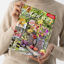 Weibliche Person im beigen Strickpullover hält ein schräg fotografiertes Magazin mit dem Titel „LandIDEE“ vor einem hellen Hintergrund. Das Cover zeigt einen blühenden Garten sowie Bildfelder mit den Aufschriften „Land erleben und genießen“, „Leckere Rezepte mit Äpfeln und Quitten“, „Herbstliche Deko für den Balkon mit Pflanzplänen“, „Wahrer Wunder-Heiler: Granatapfel“, „Bastelideen für unsere Gartentiere“ und „Leuchtfeuer für die Sinne“.