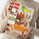 Weibliche Person im hellbeigen Strickpullover hält ein schräg ausgerichtetes Magazin mit dem Titel „Lisa Wohnen & Dekorieren“. Das Cover zeigt orange Herbstblumen, Kürbisse, Körbe und Wohnaccessoires sowie die Texte „Kreative Herbst-Ideen“, „Wohlfühl-Mix“ und „Dekolust im Herbst“ vor rotem, gelbem und grauem Hintergrund.