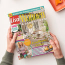 Titelblatt der Zeitschrift „Lisa Wohnen & Dekorieren“, diagonal auf weißer Fläche von zwei Händen mit grauen Strickärmeln gehalten. Das Cover zeigt ein helles Esszimmer mit Blumen, Tischdekoration und Gebäck sowie die Texte „Kreativ dem Sommer verlängern“, „Wohnen & Dekorieren“, „Landhaus-Liebe“, „EXTRA 60 Ideen für ein leichtes Wohngefühl“ und „Natürlich schön: Dekorieren mit Blumen, Gräsern & Co.“; oben rechts liegt ein orangefarbenes Gerät.
