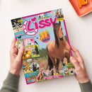 Schräg aufgenommene Zeitschrift mit pinkem Rand und blauem Cover, von zwei Händen gehalten. Sichtbare Texte: „Lissy“, „COOLER GEWINN!“, „BACKSTAGE BEIM Reitturnier“, „DIY Pferdestärke ABKÜHLUNG“, „FUN ZUM WIEHERN KOMISCH“, „Welcher PFERDE-TYP PASST zu dir?“ und „Retro Comic“; abgebildet sind mehrere Pferde, eine Reiterin und Illustrationen.