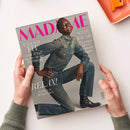 Aufgeschlagenes Magazin „MADAME“ in schräger Aufsicht, von zwei Händen über einer weißen Fläche gehalten. Das Cover zeigt eine männliche Person mit dunkler Haut in blauem Denim-Hemd und Jeans sowie schwarzem Stiefel; lesbare Texte: „WELT RÄUME“, „ENDLICH FIT“, „RELAX!“ und „Die jungen Meister des Interior-Designs“. Oben rechts liegt ein orangefarbenes Schreibutensil.