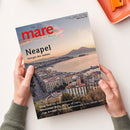 Mare-Magazin „Die Zeitschrift der Meere“ in schräger Draufsicht, von zwei Händen einer nicht näher bestimmbaren Person gehalten. Das Cover zeigt eine Küstenstadt am Meer mit Hafen, Gebäuden und Bergen bei warmem Abendlicht; sichtbare Texte: „mare“, „Neapel“, „Königin des Südens“, „Deep Talk Wie Wale telefonieren“, „Mississippi-Delta Der Blues der Cajuns“, „Cap Arcona Die vergessene Katastrophe in der Lübecker Bucht“ und „www.mare.de“. Im Hintergrund liegt ein orangefarbenes Objekt.