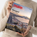 Weibliche Person im beigen Strickpullover hält eine schräg ausgerichtete Zeitschrift mit rotem Titel „mare“ und dem Untertitel „Die Zeitschrift der Meere“. Das Cover zeigt eine Küstenstadt am Meer bei Sonnenuntergang und trägt die Texte „Neapel – Königin des Südens“, „Deep Talk: Wie Wale telefonieren“, „Mississippi-Delta: Der Blues der Cajuns“ und „Cap Arcona: Die vergessene Katastrophe in der Lübecker Bucht“.