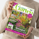 Weibliche Person im hellbeigen Strickpullover hält ein schräg ausgerichtetes Gartenmagazin mit dem Titel „Mein schöner Garten“ und dem Datum „September 2026“. Das Cover zeigt einen blühenden Garten sowie die Texte „Zucchini & Kürbis“, „Klimaschutz + Artenvielfalt“, „Kein Garten ohne Hecke“ und „Blütenglück mit pflegeleichten Fetthennen“.