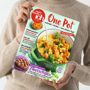 Eine Person im beigefarbenen Strickpullover hält ein schräg fotografiertes Magazin mit dem Titel „Meine Familie & Ich“ vor mintgrünem Hintergrund. Das Cover zeigt ein Gemüse-Curry mit Hähnchen, einen Kuchen und Texte wie „One Pot“, „Schnell • lecker • aus einem Topf“, „Sommerküche“, „Neue Rezepte!“ und „Traumhafte Kuchen: Zwetschgen“.