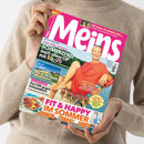 Weibliche Person im beigefarbenen Strickpullover hält eine schräg fotografierte Zeitschrift mit pinkem Titel „Meins“. Das Cover zeigt eine lächelnde Frau im roten Oberteil am Meer und trägt die Texte „Schmerzfrei auch ohne OP“, „Ab ans Meer!“, „Das hilft gegen Einsamkeit“, „Fit & Happy im Sommer“ sowie „Großes Deko-Extra“.