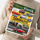Person ohne sichtbares Gesicht hält eine schräg fotografierte Zeitschrift „Motor Klassik“ vor einem hellen Hintergrund; beige Strickkleidung und zwei Hände sind erkennbar. Das Cover zeigt mehrere rote, blaue, weiße und grüne klassische Autos sowie die Texte „CABRIOLETS“, „Genießen Sie jeden Sonnenstrahl!“, „Alfa 8C Competizione“, „Mercedes 190 SL“ und das Logo „auto motor sport“.