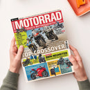 Auf weißer Fläche hält eine Person eine schräg liegende, glänzend bedruckte Zeitschrift mit rotem und weißem Titel „MOTORRAD“ und zahlreichen Motorradabbildungen. Lesbare Texte sind unter anderem „EUROPAS GRÖSSTE MOTORRADZEITSCHRIFT“, „Top-Test CFMOTO 1000 MT-X“, „BMW-NEUHEITEN“, „DUCATI PANIGALE V4/V4 S“, „KÜHLWESTEN“ und „BENELLI TRK 902“; rechts oben liegt ein orangefarbenes Tablett.