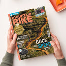 Gedrucktes Magazin „mountain BIKE“ mit orangefarbener Titelschrift, gehalten von zwei Händen einer geschlechtlich nicht erkennbaren Person über einer weißen Fläche. Das Cover zeigt einen bewaldeten Mountainbike-Trail mit fahrender Person und enthält Texte wie „E-MTB“, „Kupplungsträger“, „28 Seiten Reisen“, „4 XC-Reifen im Labor- & Praxistest“, „ROCK auf SPEED“ und „9 E-MTBs im Test“.