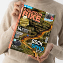 Weibliche Person im beigefarbenen Strickpullover hält ein schräg fotografiertes Magazin „mountain BIKE“ mit türkisfarbener „E-MTB“-Leiste. Das Cover zeigt einen Mountainbike-Trail, eine fahrende Person, Fahrräder auf einem „Kupplungsträger“ sowie Texte wie „28 Seiten Reise“, „14 XC-Reifen im Labor- & Praxistest“, „Eurobike kompakt“ und „Bock auf Speed“.