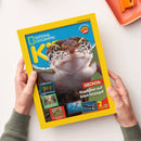 Gelbes Magazin „National Geographic“ mit großem Foto eines braun-grau gemusterten Geckos auf blauem Hintergrund, von zwei Händen einer nicht näher erkennbaren Person gehalten. Sichtbare Texte: „GECKOS: Kopfüber auf Insektenjagd!“, „2 tolle POSTER!“, „Schiffswracks: Schätze unter Wasser!“, „Sommer: Cooles über die heiße Jahreszeit“ und „Schokolade: Ihre ungewöhnliche Geschichte“. Partial orangefarbener Gegenstand oben rechts.