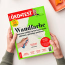 Schräg auf einem weißen Tisch liegendes Magazin „ÖKO-TEST“, gehalten von zwei Händen mit grauen Strickbündchen; roter und neongrüner Umschlag mit Porträt einer Person, weißer Farbrolle und Farbmuster. Sichtbare Texte: „Wandfarbe“, „Teuerste weiße Farbe kann nicht glänzen, günstige punkten“, „Dicke Luft?“, „Wie wir Schadstoffe in der Raumluft vermeiden“, „Test Junior-Zahnpasta“ und „Interview“.