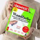 ÖKO-TEST-Magazin mit rotem Titelbalken und leuchtend grünem Rand, von einer Person im beigen Strickpullover mit beiden Händen gehalten. Auf dem Cover stehen „Wandfarbe“, „Teuerste weiße Farbe kann nicht glänzen, günstige punkten“, „Test 139 Produkte für jeden Tag“, „Dicke Luft?“ und Hinweise zu Erdbeer-Fruchtchips, Apfelessig, Fußpflege sowie Reinigungsmilch; abgebildet sind weiße Farbe und ein Pinsel.