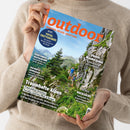 Person mit beigem Strickpullover hält ein schräg ausgerichtetes Magazin mit orangefarbenem Titel „Outdoor“ und Berglandschaft mit zwei wandernden Personen. Sichtbare Texte: „Wandern. Reisen. Abenteuer.“, „Total trittsicher!“, „Traumhafte Alpen“, „Japans Wanderjuwel“, „Quer durch Deutschland“ und „Kochen & Genießen“.