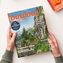 Schräg auf weißem Untergrund liegendes Magazin „outdoor“, von zwei Händen gehalten, mit glänzendem Coverfoto einer alpinen Felslandschaft, grünen Bergen und zwei wandernden Personen. Sichtbare Texte: „WANDERN. REISEN. ABENTEUER.“, „Traumhafte Alpen“, „Japans Wanderjuwel“, „Kochen & Genießen“, „Rucksäcke“ und „TOTAL TRITTSICHER!“.