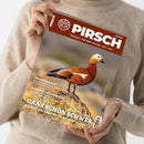 Weibliche Person im beigefarbenen Strickpullover hält schräg eine Zeitschrift mit braunem Titelbereich. Sichtbar sind das Logo „PIRSCH“, „RESPEKT VOR DEM WILDEN. SEIT 1879.“, eine Rostgans sowie die Überschriften „WASCHBÄREN“, „AUFBRUCHSSTIMMUNG“ und „GÄNSE SCHÖN SCHWER“.