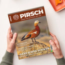 Schräg gehaltenes PIRSCH-Magazin in zwei Händen einer nicht näher bestimmbaren Person, mit dem Titel „PIRSCH“, dem Text „RESPEKT VOR DEM WILDEN. SEIT 1879.“ und dem Foto einer braun-orangefarbenen Gans auf einem Felsen. Sichtbare Überschriften: „WASCHBÄREN“, „AUFBRUCHSTIMMUNG“ und „GANS SCHÖN SCHWER“; oben rechts liegt ein orangefarbenes Etui.