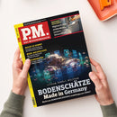 Schräg gehaltenes schwarzes PM-Magazin mit gelbem Rand, rotem Logo „PM. DAS WISSENSMAGAZIN“ und zwei Händen in grauen Ärmeln vor weißem Hintergrund. Das Cover zeigt Gesteinsproben mit Leuchtschildern chemischer Elemente, ein Toilettenfoto sowie die Texte „BODENSCHÄTZE Made in Germany“, „LITHIUM, KOBALT, WOLFRAM“, „GROSSES GESCHÄFT Hightech-Klos analysieren unsere Gesundheit“, „MACHT KI DUMM?“ und „WEIN-ARCHÄOLOGIE“.