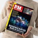 Weibliche Person in beigefarbenem Strickpullover hält ein schräg abgebildetes Magazin mit schwarzem Cover und gelbem Rand. Sichtbare Texte: „PM! DAS WISSENSMAGAZIN“, „MACHT KI DUMM?“, „WEIN-ARCHÄOLOGIE“, „LÖSCHROBOTER“, „GROSSES GESCHÄFT: Hightech-Klos analysieren unsere Gesundheit“ und „BODENSCHÄTZE – Made in Germany“.