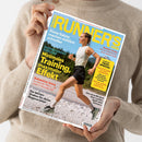 Schräg gehaltenes Runners-World-Laufmagazin mit weißem Rand und blau-gelbem Titel „RUNNERS WORLD“, darunter „Das größte Laufmagazin der Welt“. Auf dem Cover läuft eine weibliche Person in weißem Oberteil, schwarzer Hose und Laufschuhen; weitere Texte lauten „Diese Salate machen wirklich schneller“, „Minimales Training, maximaler Effekt“, „Stabiler laufen“ und „Trail-Special“. Das Magazin wird von einer Person im beigefarbenen Strickpullover gehalten.