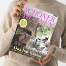Person mit beigefarbenem Strickpullover hält ein schräg fotografiertes Wohnmagazin mit grauem Cover und pinkem Titel „SCHÖNER WOHNEN“. Sichtbar sind „Europas größtes Wohnmagazin“, „Facelift für die Küche“, „Raumwunder“, „Überraschung!“, „Beton, Holz, Farbe“ sowie „Das hat Klasse“; das Cover zeigt ein Wohnzimmer mit Kamin, Sofa, Sesseln und Gartenblick.