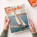 Auf weißem Untergrund hält eine nicht näher erkennbare Person mit beiden Händen das Magazin „segeln“ im Hochformat. Das Cover zeigt ein weißes Segelboot auf blauem Meer und trägt die Texte „Juli August 2026“, „Fahrten- und Freizeitsegeln“, „Nach Gefühl … Segeln ohne Technik“, „Recht & Technik“, „Dänemark – Fünen und Seeland“ sowie „8 SEITEN GEBRAUCHTBOOTE“; oben rechts liegt ein orangefarbenes Notizbuch.