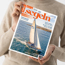 Weibliche Person im beigen Strickpullover hält schräg eine Zeitschrift mit orangefarbenem Titel „segeln“ und dem Hinweis „Fahrten- und Freizeitseglen“. Das Cover zeigt ein weißes Segelboot auf blauem Meer sowie die Texte „Dänemark – Fünen und Seeland“, „Deckssalon im Eigenbau“, „Nach Gefühl … Segeln ohne Technik“ und „8 Seiten Gebrauchtboote“.