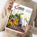 Weibliche Person im beigefarbenen Strickpullover hält schräg eine weiße Zeitschrift mit dem Titel „SENUS“ und dem Untertitel „einfach gut leben“. Das Cover zeigt bunte Blumen, eine gelbe Kanne und gerollte helle Tücher auf einem Holztisch sowie die Texte „Der beste Kaiserschmarrn der Welt“, „BALSAM FÜR DIE SEELE“ und „Wie uns die Natur mehr Lebensfreude schenkt“.