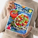 Eine Person im beigefarbenen Strickpullover hält schräg ein Kochmagazin mit dem Titel „So isst Italien“. Das Cover zeigt einen bunten Tomaten-Mozzarella-Salat, die Texte „94 geniale Rezepte“, „30 x schnell und einfach kochen“, „Italienische Urlaubsküche“, „Pomodori!“ und „Dolce vita auf dem Teller!“ sowie eine Frau im roten Trägerkleid.