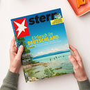 Aufgeschlagenes „stern“-Magazin mit blauem Cover, gelber Überschrift „Urlaub in DEUTSCHLAND“ und Titelfoto eines türkisfarbenen Sees mit Bergen, Ufer und badenden Menschen. Zwei Hände mit grauen Strickärmeln halten das Magazin; sichtbar sind außerdem „FUSSBALL WM 2026“ und „Die große WM-Bilanz: Wer die Herzen gewann, welche Pannen passierten, wie die Fifa die Zukunft plant“.
