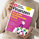Weibliche Person im beigefarbenen Strickpullover hält ein schräg fotografiertes Magazin mit dem Titel „Stiftung Warentest Finanztest“. Das magentafarbene Cover trägt den Text „Der große Versicherungs-Check“, zeigt eine stilisierte Person mit Fernrohr auf bunten Blöcken und einen gelben Hinweis „Frist für Steuererklärung“.
