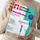 Person mit beigefarbenem, grob gestricktem Pullover hält eine schräg ausgerichtete Zeitschrift aus bedrucktem Papier; beide Hände und ein abgebildeter blauer Farbroller sind sichtbar. Das türkis-weiße Cover trägt die Aufschrift „Stiftung Warentest“, den Titel „Wandfarben“ sowie Texte zu Kinderwagen, Geschirrspültabs, Druckern und „Diagnose per KI“.