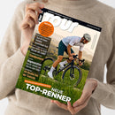 Diagonal gehaltenes, glänzendes TOUR-Rennradmagazin mit schwarzem Rand und großem weißem Titel „TOUR“, Aufschrift „EUROPAS RENNRAD-MAGAZIN NR. 1“ und „www.tour-magazin.de“. Das Cover zeigt einen männlichen Rennradfahrer mit weißem Helm auf schwarzem Fahrrad vor grüner Landschaft; eine weibliche Person im beigefarbenen Strickpullover hält das Magazin mit beiden Händen.