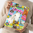 Weibliche Person im beigen Strickpullover hält eine schräg fotografierte, farbig bedruckte Pferdezeitschrift. Das Cover zeigt ein weißes Pferd in der Mitte, ein braunes Pferd rechts und mehrere kleinere Pferdefotos; lesbare Texte sind „5 Sommer POSTER Pferde für KÖNIGE“, „SAFE IM SATTEL!“, „SPASS am Strand“ und „BRAVO“.