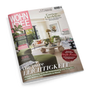 Schräg fotografiertes Cover der Wohnzeitschrift „WOHNIDEE“ aus glänzendem Papier vor schwarzem Hintergrund, mit einem hell eingerichteten Wohnzimmer, grüner Pflanze, Sofa, Couchtisch, Sesseln, Lampe, Bildern und Dekoration. Sichtbare Texte: „Garten-Checkliste“, „Sommer bewahren“, „BITTE ZU TISCH“, „KÜHLE BRISE“, „Der große Sofa-Guide“ und „Die pure LEICHTIGKEIT“.